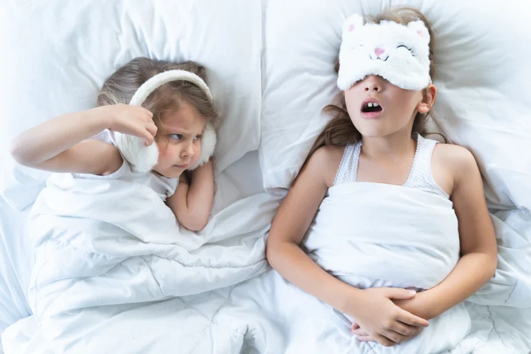 Two girls in bed, one snoring with her mouth open while her sister covers her ears